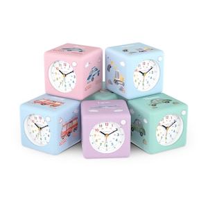 Könnte beinhalten: Eine Sammlung von fünf würfelförmigen Kinderweckern in Pastellfarben. Jede Uhr hat ein weißes Zifferblatt mit schwarzen Zeigern und bunten Zahlen. Die Uhren sind mit Illustrationen von Fahrzeugen verziert, darunter ein Polizeiauto, ein Feuerwehrauto und ein Flugzeug.