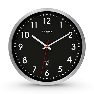 Puede incluir: Un reloj de pared negro y plateado con números blancos y una aguja de segundos roja. El reloj es de radiocontrol y tiene el texto "CANDER BERLIN" y "FUNKGESTEUERT" impreso en la esfera.