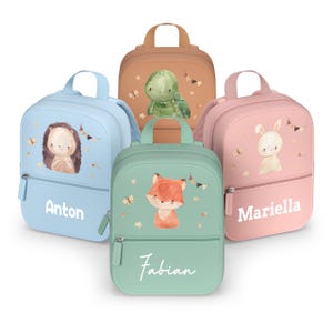 Pode incluir: Cinco mochilas coloridas com ilustrações de animais e nomes personalizados. As mochilas são azul claro, castanho, rosa e verde. Cada mochila apresenta um animal diferente: um ouriço, uma tartaruga, um coelho e uma raposa. Os nomes "Anton", "Fabian" e "Mariella" estão impressos.