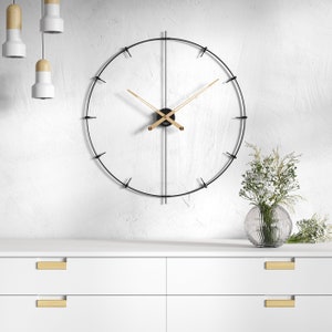 Könnte beinhalten: Eine moderne, minimalistische Wanduhr mit einem schwarzen Metallrahmen und goldfarbenen Zeigern. Die Uhr hat ein schlichtes, elegantes Design mit einer kreisförmigen Form und ist an einer weißen Wand befestigt. Die Uhr hat einen Durchmesser von etwa 61 cm.