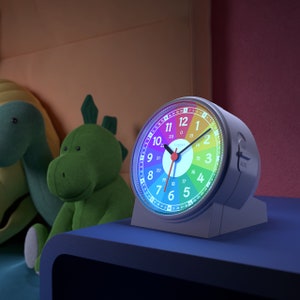 Op de afbeelding: Een witte analoge wekker met een regenboogkleurig gezicht en zwarte wijzers. De wijzerplaat van de klok heeft de nummers 1-12 en 1-24 rond de rand. De klok staat op een blauw oppervlak.