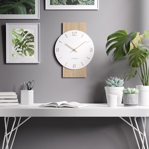 Könnte beinhalten: Eine moderne Wanduhr mit weißem Zifferblatt und goldfarbenen Zeigern und Ziffern. Die Uhr ist auf einer rechteckigen Holzrückwand montiert. Das Wort "Cander" ist darunter aufgedruckt. Umgeben von gerahmten botanischen Drucken und Topfpflanzen.