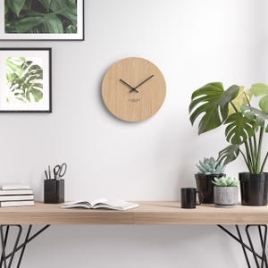 Könnte beinhalten: Eine runde Wanduhr aus hellem Holz mit schwarzen Zeigern und minimalistischen Stundenmarkierungen. Die Uhr, mit der Marke "Cander", ist an einer weißen Wand befestigt. Ein Holztisch darunter trägt eine schwarze Tasse, Pflanzen und Bücher, für einen modernen Look.