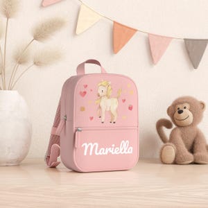 Peut inclure: Un sac à dos rose orné d'une illustration de poney, de petits cœurs rouges et de points jaunes sur la poche avant. Le prénom Mariella est inscrit en lettres cursives blanches sous le poney. Le sac possède une poignée et des bretelles réglables.