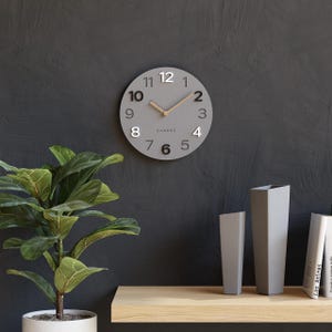 Könnte beinhalten: Eine graue runde Wanduhr mit schwarzen Ziffern und Holzzeigern hängt an einer dunkelgrauen Wand. Auf dem Zifferblatt steht das Wort "Cander". Eine Topfpflanze und Vasen stehen auf einem Holzregal darunter.