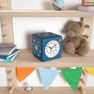Könnte beinhalten: Eine blaue, würfelförmige Uhr mit weißem Zifferblatt und orangefarbener Taste. Die Uhr hat ein Weltraum-Design mit Planeten und Sternen. Ein Teddybär und Bücher befinden sich auf dem Regal. Die Uhr misst etwa 10 cm pro Seite.