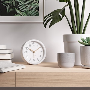 May include: A white analog clock with a round face, featuring black numerals and gold-colored hands. The clock is on a wooden surface, with a stack of books and a fern print in the background. The clock's face reads "Cander".