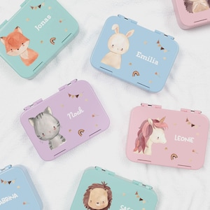 Könnte beinhalten: Eine Sammlung personalisierter Metall-Lunchboxen in verschiedenen Pastellfarben. Jede Box zeigt eine andere Tierillustration, darunter ein Fuchs, ein Hase, eine Katze und ein Einhorn, mit einem auf dem Deckel aufgedruckten Namen. Dekorative Elemente sind Regenbogen und Wimpelketten.