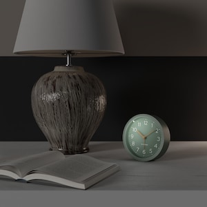 May include: A bedside scene featuring a green analog alarm clock with a light wood-colored hand, a ceramic lamp with a gray lampshade, and an open book. The clock is round and has white numbers.