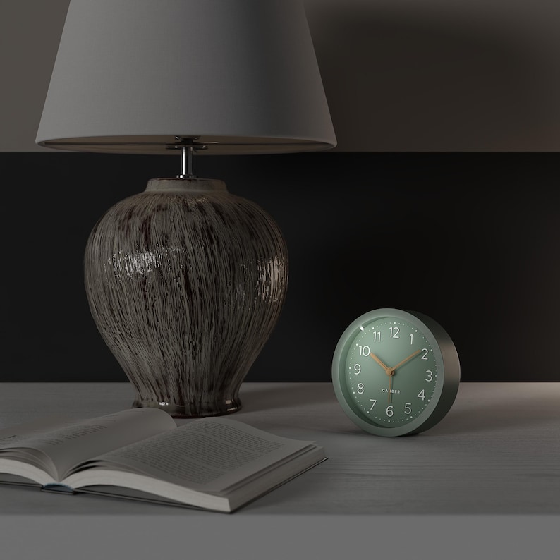 May include: A bedside scene featuring a green analog alarm clock with a light wood-colored hand, a ceramic lamp with a gray lampshade, and an open book. The clock is round and has white numbers.