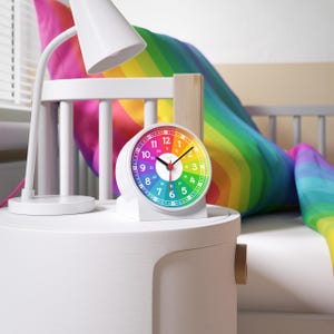 May include: A white alarm clock with a rainbow-colored face sits on a white nightstand. The clock has black hands and a red second hand. A white lamp is to the left of the clock. A rainbow-colored pillow is in the background.