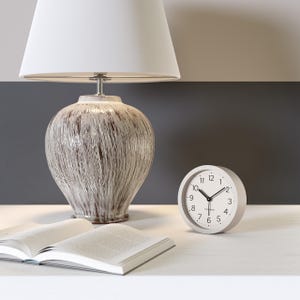Könnte beinhalten: Eine weiße Tischlampe mit weißem Lampenschirm und einem strukturierten, braun-weißen Keramikfuß. Eine kleine, runde, graue Analoguhr und ein offenes Buch liegen auf dem Tisch. Die Uhr zeigt die Uhrzeit an.