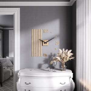 Può includere: Un orologio da parete moderno con quadrante quadrato e lancette nere. L'orologio presenta un pannello in legno con doghe verticali e numeri individuali in legno. L'orologio è montato su una parete grigia sopra un comò bianco.