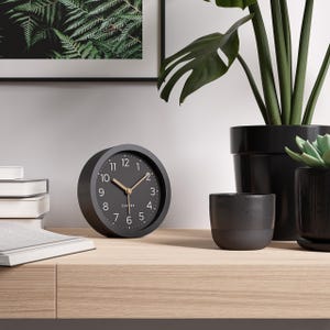 Könnte beinhalten: Eine schwarze analoge Uhr mit goldenen Zeigern und weißen Ziffern. Das Zifferblatt hat ein minimalistisches Design. Das Wort "CAMER" ist unten auf dem Zifferblatt aufgedruckt. Die Uhr steht auf einer Holzoberfläche.