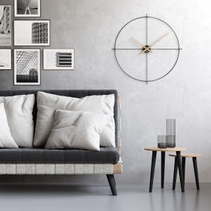 Op de afbeelding: Een moderne woonkamer met een donkergrijze bank met witte kussens, een minimalistische zwarte wandklok met houten wijzers en twee kleine houten bijzettafels met zwarte poten. Verschillende ingelijste zwart-wit kunstwerken hangen aan de grijze muur.