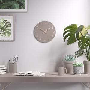 Pode incluir: Um relógio de parede redondo, cor de taupe, com um design minimalista. O relógio apresenta um mostrador claro com algarismos e ponteiros brancos simples. A palavra "CAMDEN" está impressa abaixo do centro. O relógio está montado numa parede branca, com uma secretária e plantas em primeiro plano.