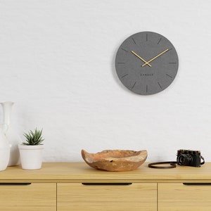 Può includere: Un orologio da parete rotondo grigio con lancette dorate e la scritta "CANDER" sul quadrante. L'orologio è montato su una parete bianca sopra un mobile in legno. Una piccola pianta grassa in un vaso bianco, una ciotola di legno e una fotocamera vintage sono sul mobile.