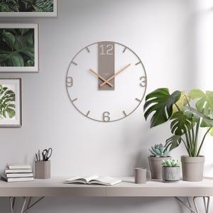Peut inclure: Une horloge murale moderne au design minimaliste. L'horloge présente un cadran transparent avec un cadre et des aiguilles gris, et un accent rectangulaire gris en haut avec le chiffre 12. L'horloge est fixée sur un mur blanc au-dessus d'un bureau avec des plantes et de la décoration.