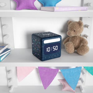 Könnte beinhalten: Ein marineblauer digitaler Wecker mit Sternenmuster und der angezeigten Uhrzeit 10:50. Ein kleiner Teddybär sitzt in der Nähe auf einem weißen Regal, zusammen mit einem Buch und sternförmigen Kissen. Ein farbenfrohes Banner hängt darunter.