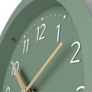 May include: Close-up of a sage green analog clock. The clock face features white numerals and small dots marking the minutes. The hands are made of light-colored wood. The clock is a simple, modern design.