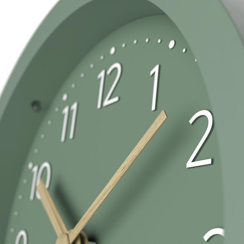 May include: Close-up of a sage green analog clock. The clock face features white numerals and small dots marking the minutes. The hands are made of light-colored wood. The clock is a simple, modern design.