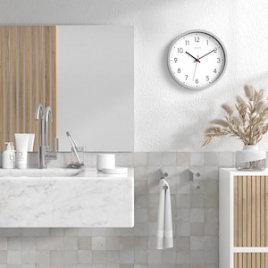 Könnte beinhalten: Ein modernes Badezimmer mit einem weißen Marmorwaschbecken, einer silbernen Armatur und einer runden Wanduhr mit grauem Rahmen. Ein weißes Handtuch hängt an einem silbernen Haken. Eine Vase mit getrockneten Pflanzen steht auf einem weißen Schrank.
