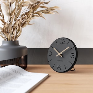Op de afbeelding: Een grijze analoge klok met zwarte cijfers en wijzers, en het woord "CANDER" gedrukt op de wijzerplaat. De klok staat op een houten oppervlak, met een vaas met gedroogde planten en een open boek op de achtergrond.