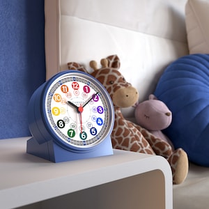 Peut inclure: Un réveil analogique bleu avec un cadran coloré affichant des chiffres et une trotteuse rouge. La montre est posée sur une table de chevet blanche, avec une girafe et un animal en peluche rose en arrière-plan. Le cadran de l'horloge porte le mot "CANDER".