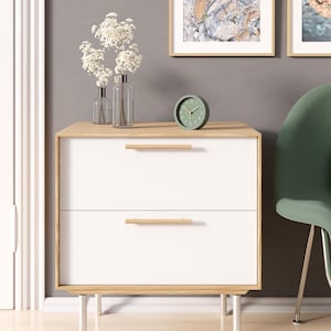 May include: A modern white nightstand with two drawers and light wood accents. The nightstand has a light wood top and drawer pulls. A small green clock and vases with white flowers sit on top. The nightstand is on silver legs.