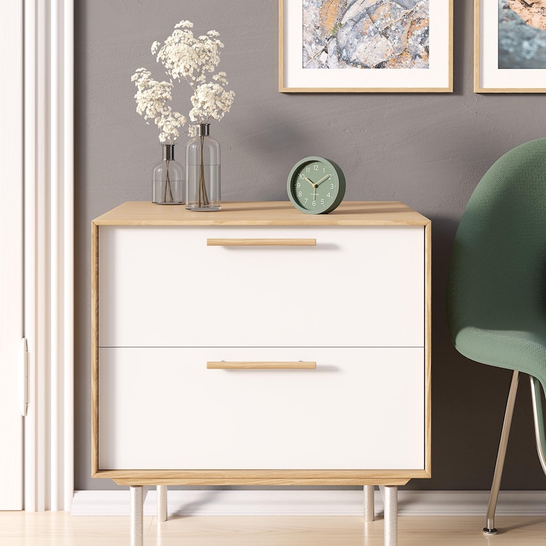 May include: A modern white nightstand with two drawers and light wood accents. The nightstand has a light wood top and drawer pulls. A small green clock and vases with white flowers sit on top. The nightstand is on silver legs.