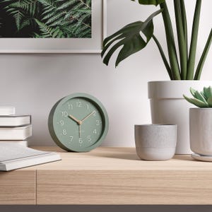 May include: A sage green analog clock with a round face, featuring white numerals and hands. The clock is approximately 4 inches in diameter. It sits on a light wood surface, near potted plants and a framed fern print.