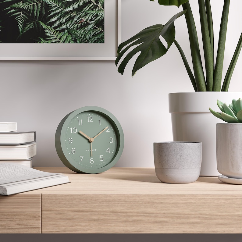 May include: A sage green analog clock with a round face, featuring white numerals and hands. The clock is approximately 4 inches in diameter. It sits on a light wood surface, near potted plants and a framed fern print.