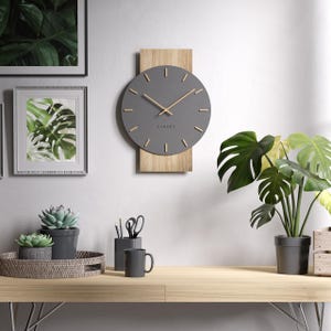 Puede incluir: Un moderno reloj de pared gris con detalles en madera. La esfera del reloj es redonda con marcadores de hora y manecillas minimalistas. La palabra "CANDER" está impresa debajo del centro. El reloj está montado en un panel de madera clara. La imagen también incluye un escritorio con plantas y decoración.