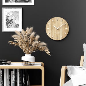 May include: A wooden wall clock with white hands and markers hangs on a dark wall. A white vase with dried flowers sits on a wooden table. A framed black and white botanical print is visible.