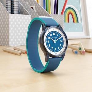 Op de afbeelding: Een blauw en turquoise horloge met een ronde wijzerplaat en een blauwe stoffen band. De wijzerplaat heeft witte cijfers en wijzers, met de merknaam "CANDER". Het horloge ligt op een houten oppervlak, met potloden en bureau-organizers op de achtergrond.