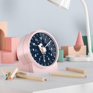 Könnte beinhalten: Ein rosa, runder Wecker mit einem dunkelblauen Zifferblatt mit einer weißen Einhorn-Illustration. Die Uhr hat weiße Zeiger und einen weißen Rand. Die Uhr steht auf einer weißen Oberfläche mit Holzstiften und anderen dekorativen Gegenständen.