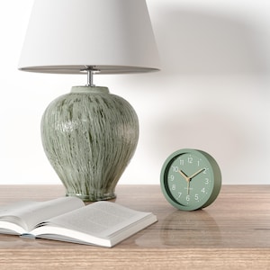 Könnte beinhalten: Eine salbeigrüne Tischlampe mit weißem Lampenschirm steht neben einer kleinen, runden, grünen Uhr. Ein offenes Buch liegt auf einer Holzoberfläche. Die Uhr hat goldene Zeiger und weiße Zahlen.