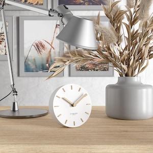 Könnte beinhalten: Eine weiße, runde Uhr mit goldfarbenen Zeigern und Markierungen, der Text "CANDER BERLIN" ist auf dem Zifferblatt aufgedruckt. Die Uhr steht auf einer Holzoberfläche neben einer grauen Vase mit getrockneten Pflanzen und einer silbernen Schreibtischlampe.