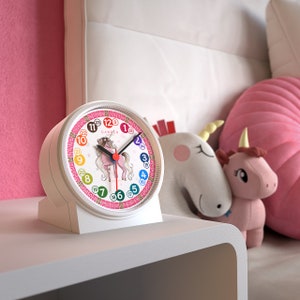 Könnte beinhalten: Weißer Wecker mit einem rosa Einhorn-Design. Das Zifferblatt hat bunte Zahlen und den Markennamen "CANDER". Die Uhr steht auf einem weißen Nachttisch, mit Einhorn-Plüschtieren im Hintergrund.