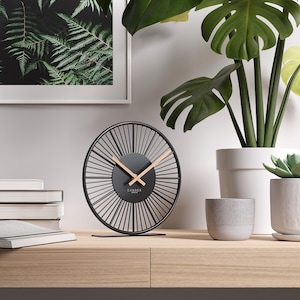Op de afbeelding: Een zwarte en gouden analoge klok met een radiaal ontwerp, met een zwarte wijzerplaat en gouden wijzers. De klok staat op een houten oppervlak, met een ingelijste varenprint en potplanten op de achtergrond. De klok leest "Cander Berlin".