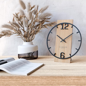 Könnte beinhalten: Eine moderne Holz-Tischuhr mit schwarzem Metallrahmen und Zeigern. Das Zifferblatt zeigt die Zahlen 12 und 6, darunter steht der Markenname "CANDER BERLIN". Die Uhr steht auf einer Holzoberfläche, neben einer Vase mit getrockneten Pflanzen.