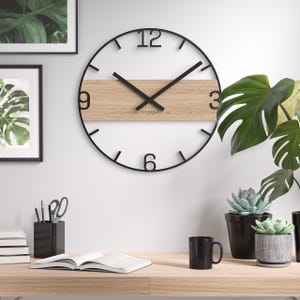 Op de afbeelding: Een moderne wandklok met een zwart metalen frame en wijzers. De wijzerplaat heeft een licht houten paneel in het midden met de cijfers 3, 6, 9 en 12 in zwart. De klok staat op een witte muur met ingelijste kunstwerken en planten.