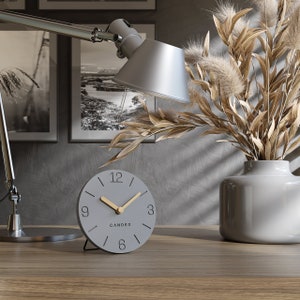 Può includere: Un orologio analogico grigio con lancette dorate e un'iscrizione dorata che recita "CANDER" su un tavolo di legno grigio. L'orologio è davanti a un vaso con erba della pampa secca.