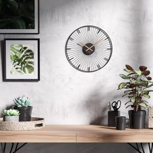 Könnte beinhalten: Eine schwarze Metall-Wanduhr mit radialem Design und goldfarbenen Zeigern. Die Uhr ist an einer weiß strukturierten Wand angebracht, mit gerahmten botanischen Drucken und Topfpflanzen auf einem Holzschreibtisch. Der Durchmesser der Uhr beträgt ca. 46 cm.