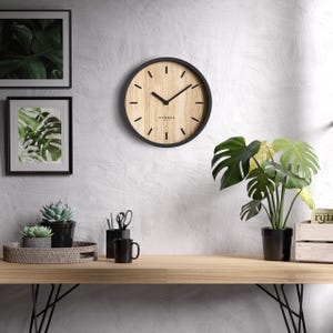 Könnte beinhalten: Eine runde Wanduhr mit schwarzem Rahmen und hellem Holzmaserungszifferblatt. Die Uhr hat schwarze Zeiger und minimalistische Stundenmarkierungen. Das Wort "Cander" ist auf dem Zifferblatt aufgedruckt. Die Uhr ist an einer weißen Wand mit gerahmten botanischen Drucken befestigt.