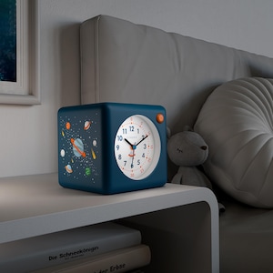 Könnte beinhalten: Ein marineblauer, würfelförmiger Wecker mit weißem Zifferblatt und orangefarbener Taste. Das Zifferblatt hat schwarze Zahlen und Zeiger. Die Seite der Uhr zeigt ein Weltraum-Design mit Planeten und Sternen. Die Uhr steht auf einem Nachttisch.