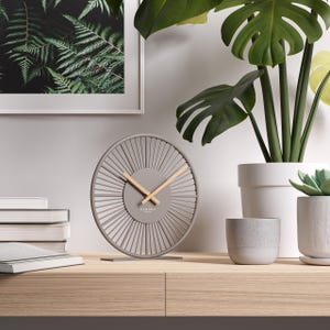 Op de afbeelding: Een grijze en houten analoge klok met een radiaal ontwerp, houten wijzers en het woord "Cander" op de wijzerplaat. De klok staat op een houten oppervlak, naast potplanten en een ingelijste varenprint.