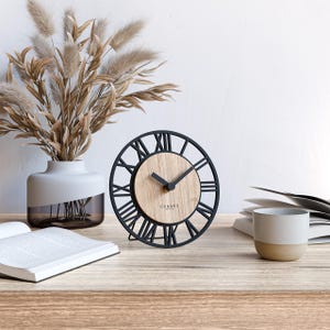 Può includere: Un orologio rotondo con una cornice in metallo nero e numeri romani. Il quadrante è in legno chiaro e le lancette sono nere. L'orologio è su una superficie di legno, con un vaso di fiori secchi e una tazza di caffè nelle vicinanze.