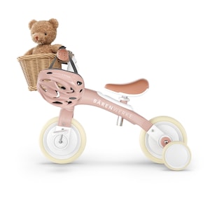 Op de afbeelding: Een roze loopfiets met crèmekleurige wielen en een bruin zadel. Een rieten mand op het stuur bevat een teddybeer. Een bijpassende roze helm is bevestigd. De tekst "BÄRENWERKE" staat op het frame.
