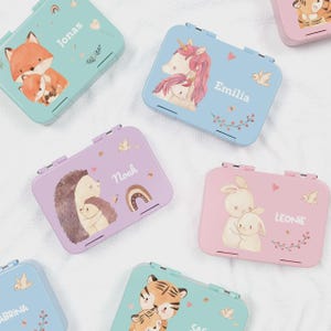 Könnte beinhalten: Eine Sammlung pastellfarbener rechteckiger Metall-Lunchboxen. Jede Box zeigt eine andere Tierillustration, darunter ein Fuchs, ein Einhorn, ein Igel und ein Hase, mit dem Namen eines Kindes auf dem Deckel. Die Deckel haben einen Klappverschluss.